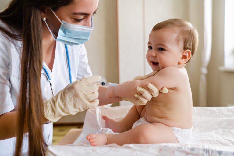 Young female pediatrician or nurse giving an intramuscular injection of a vaccine to hand of little baby boy Immunization for children concept. Happy little cute boy getting a flu shot not afraid of the syringe needle